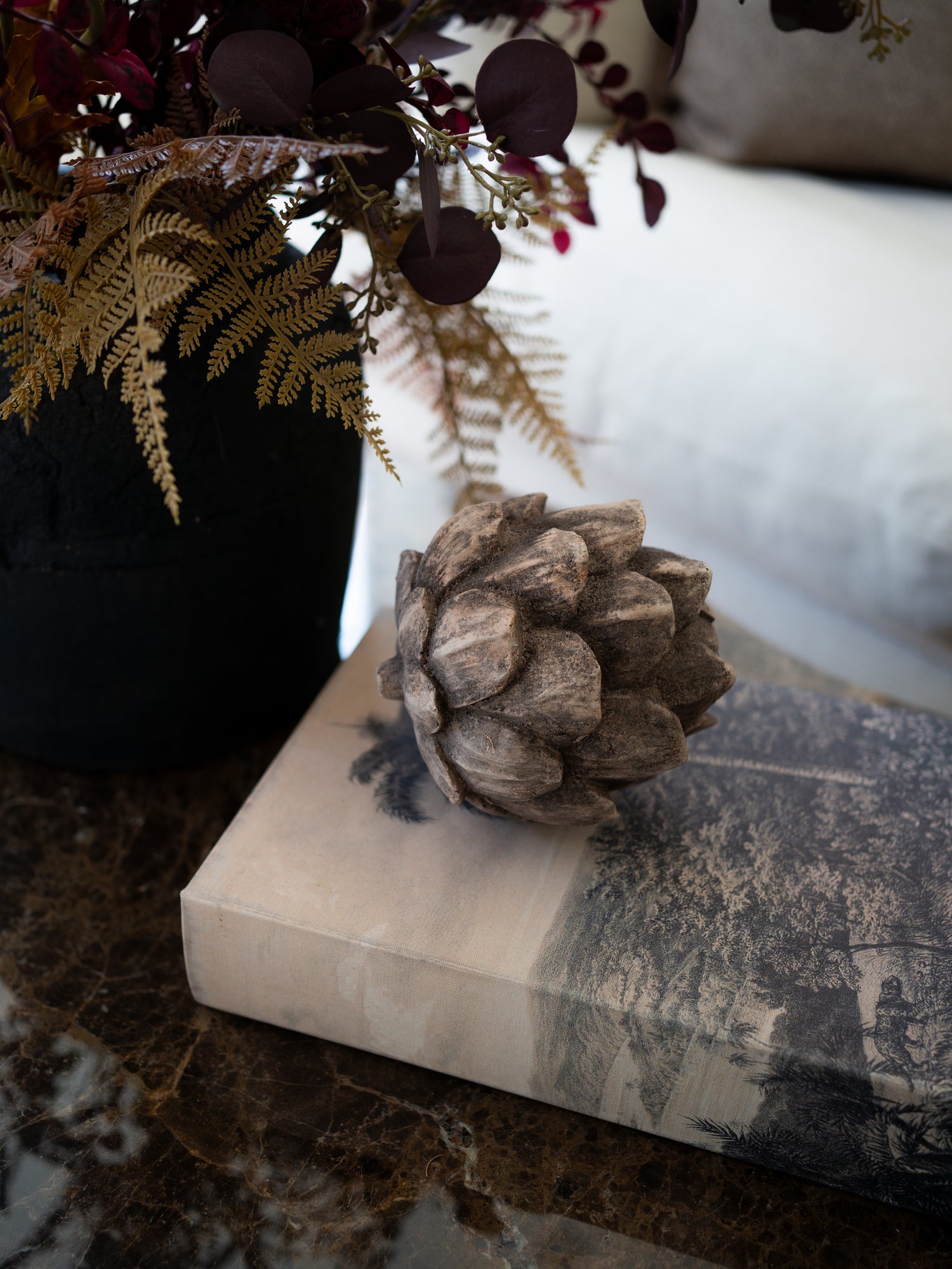 A decorative rustic pinecone-style decor piece resting on a beige book with a black-and-white landscape print, set on a dark marble surface next to a black vase with dried foliage.