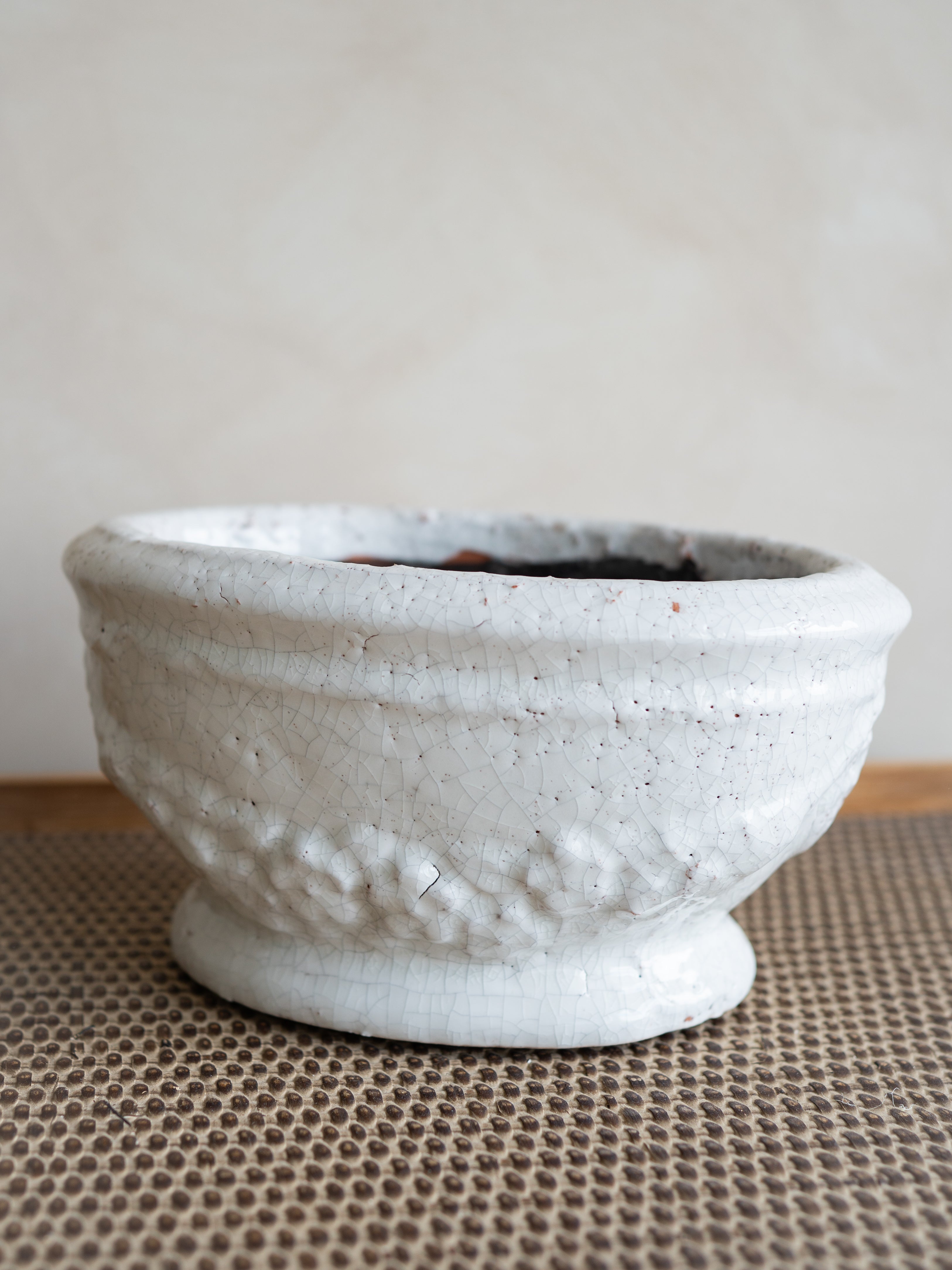 A small handmade ceramic plant pot with a textured, hammered surface and a footed base, finished in a matte off-white glaze, resting on a brown woven mat against a neutral beige background.