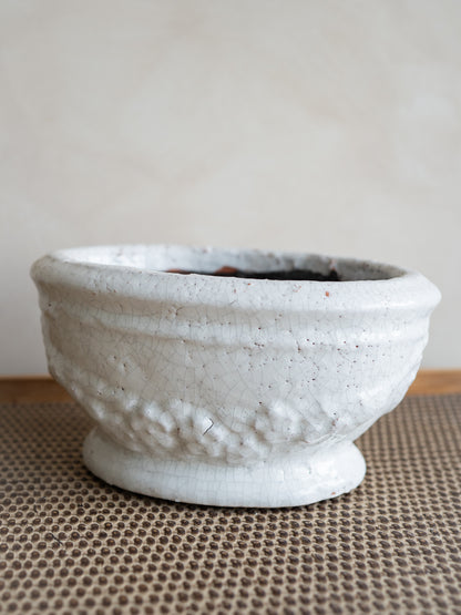 A small handmade ceramic plant pot with a textured, hammered surface and a footed base, finished in a matte off-white glaze, resting on a brown woven mat against a neutral beige background.