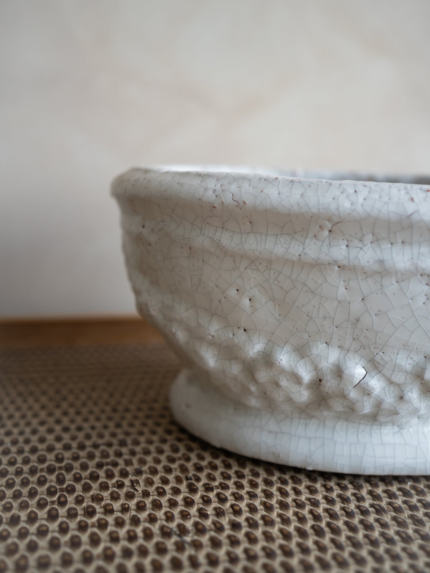 A close-up of a textured ceramic planter with a white glaze and crackle pattern, resting on a brown woven mat against a neutral beige background.