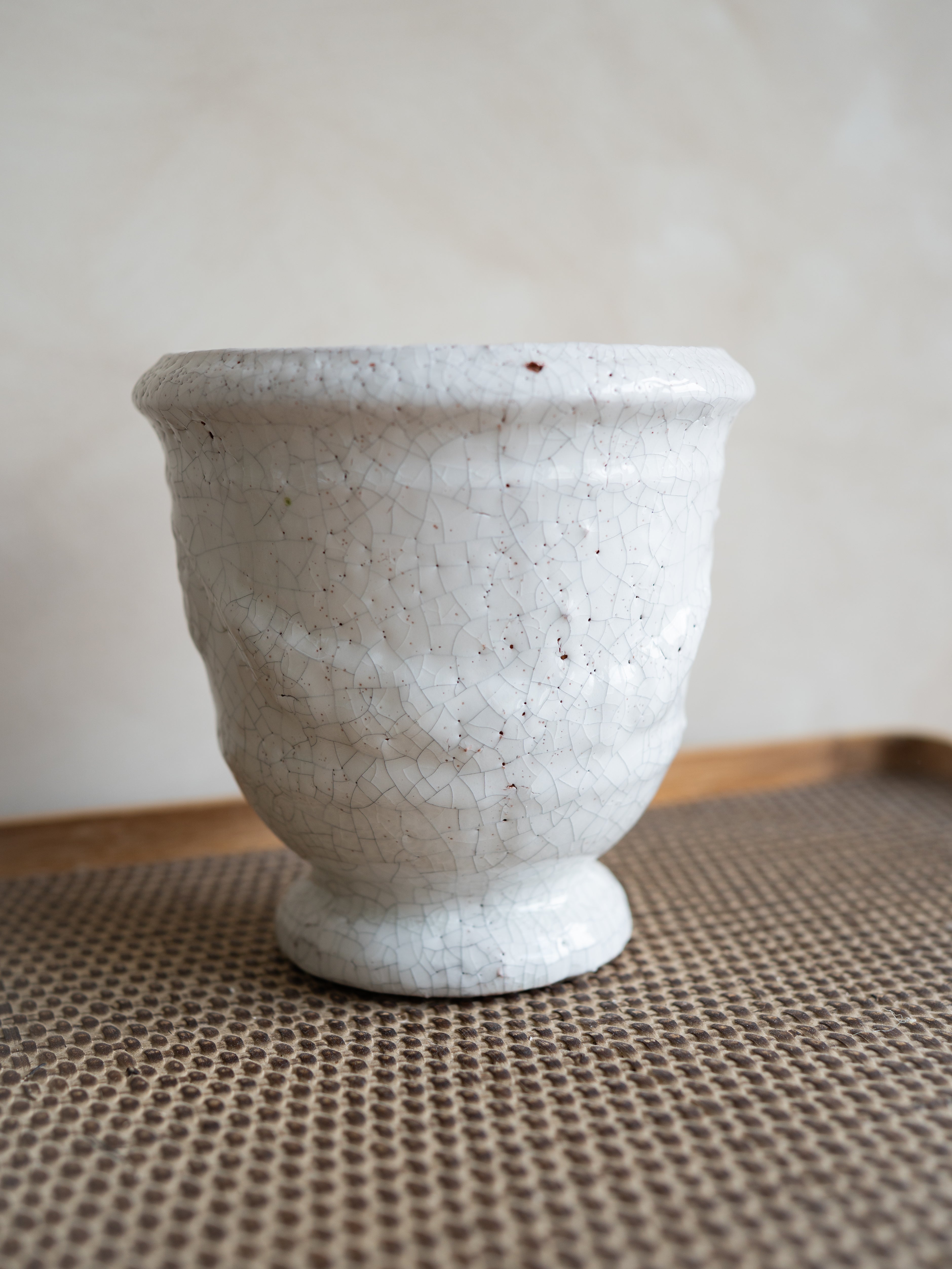 A small white ceramic planter with a textured, crackled glaze and a flared rim, sitting on a brown woven mat against a neutral beige background.