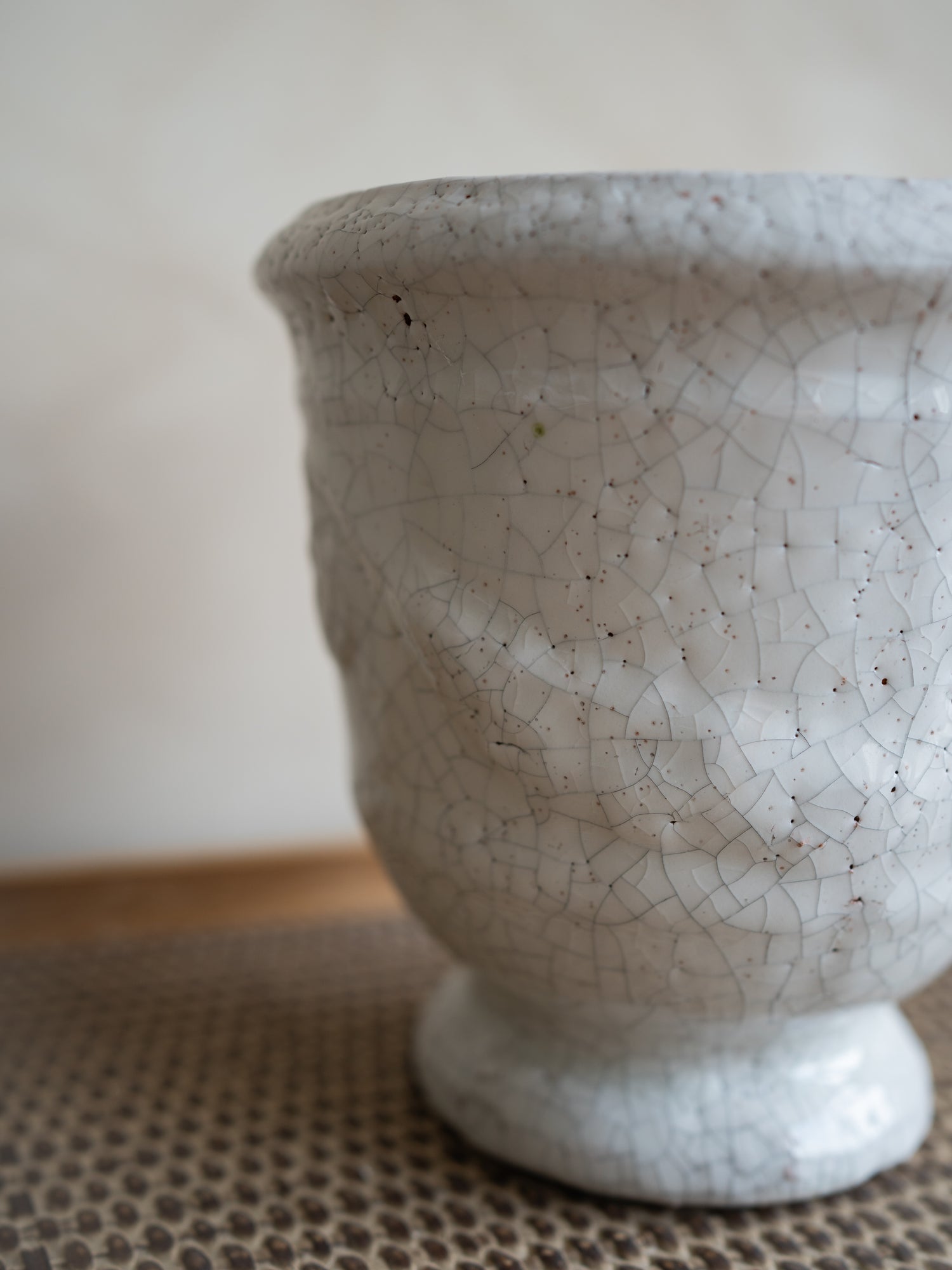 A close-up of a ceramic planter pot with a textured, footed base and a white glaze featuring a crackle pattern with dark speckles, resting on a woven mat.