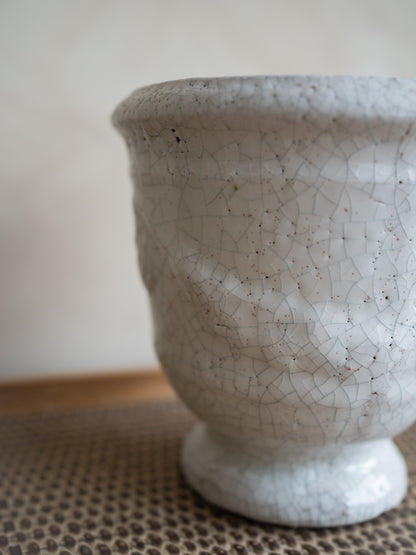 A close-up of a ceramic planter pot with a textured, footed base and a white glaze featuring a crackle pattern with dark speckles, resting on a woven mat.