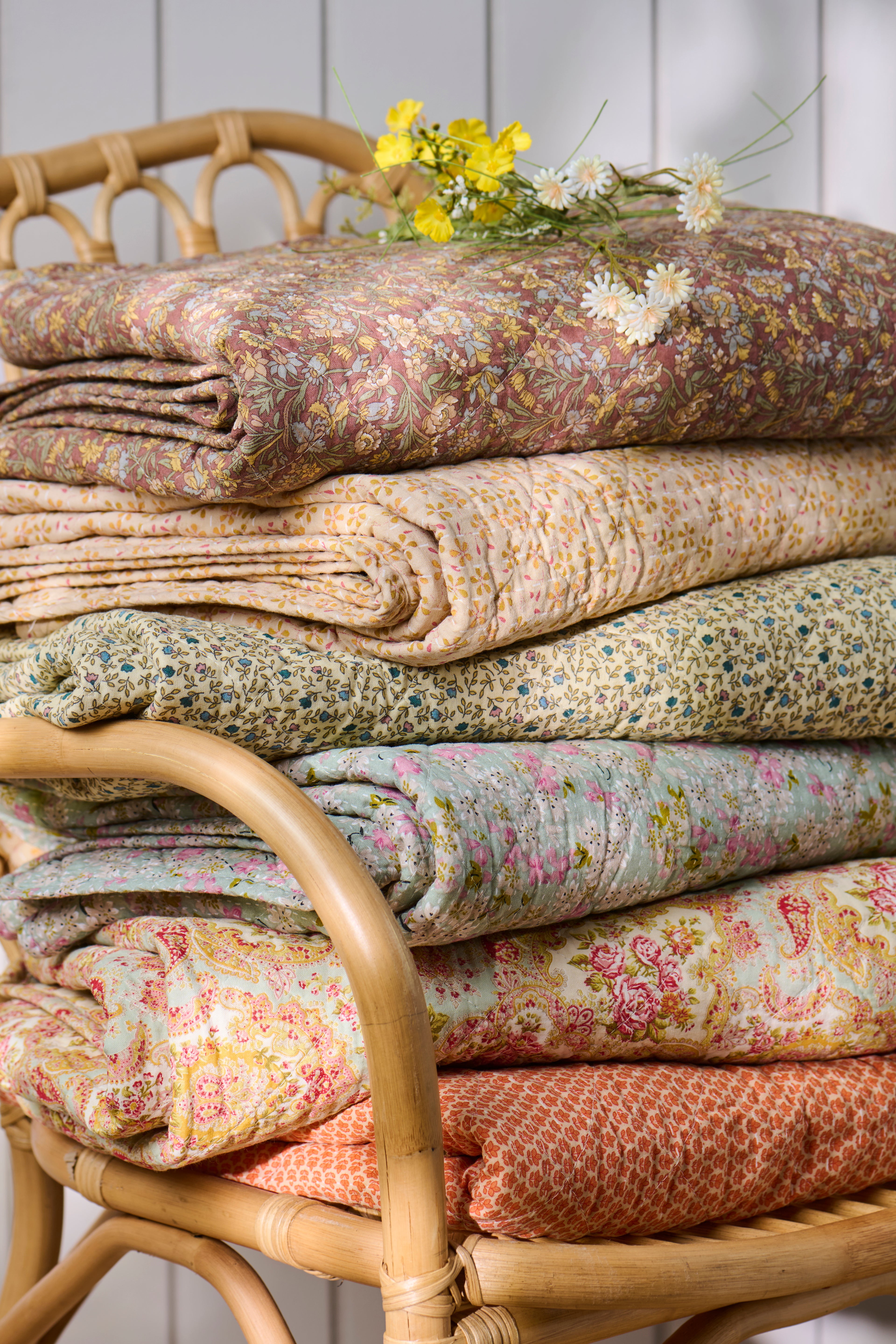 Stack of floral-patterned fabrics on a wicker chair with a white wooden panel background