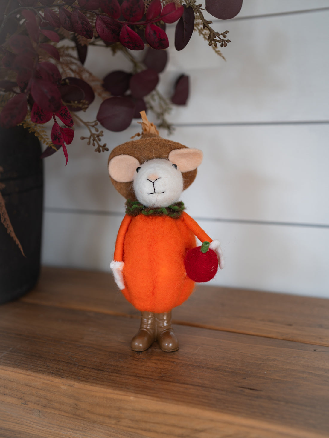 A small felt pumpkin character figurine wearing a brown hat and holding a red apple, displayed on a wooden surface against a white paneled wall with dried foliage in the background.