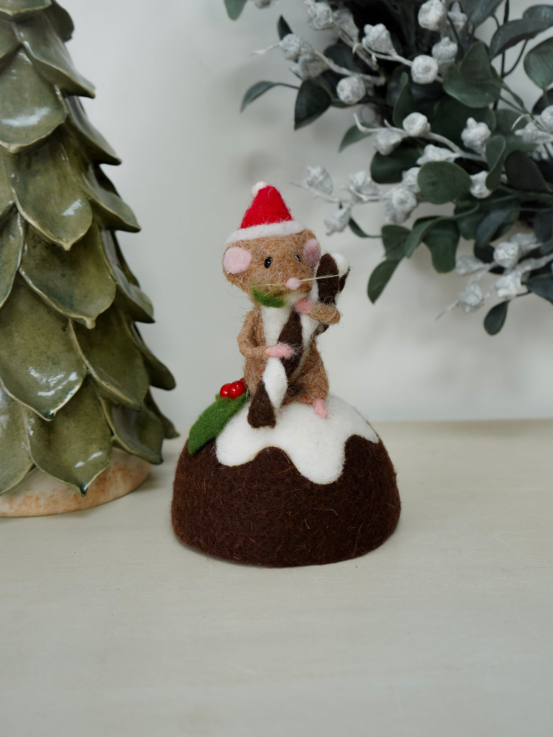 Decorative mouse in a Santa hat on a Christmas pudding with a ceramic tree and mistletoe in the background.
