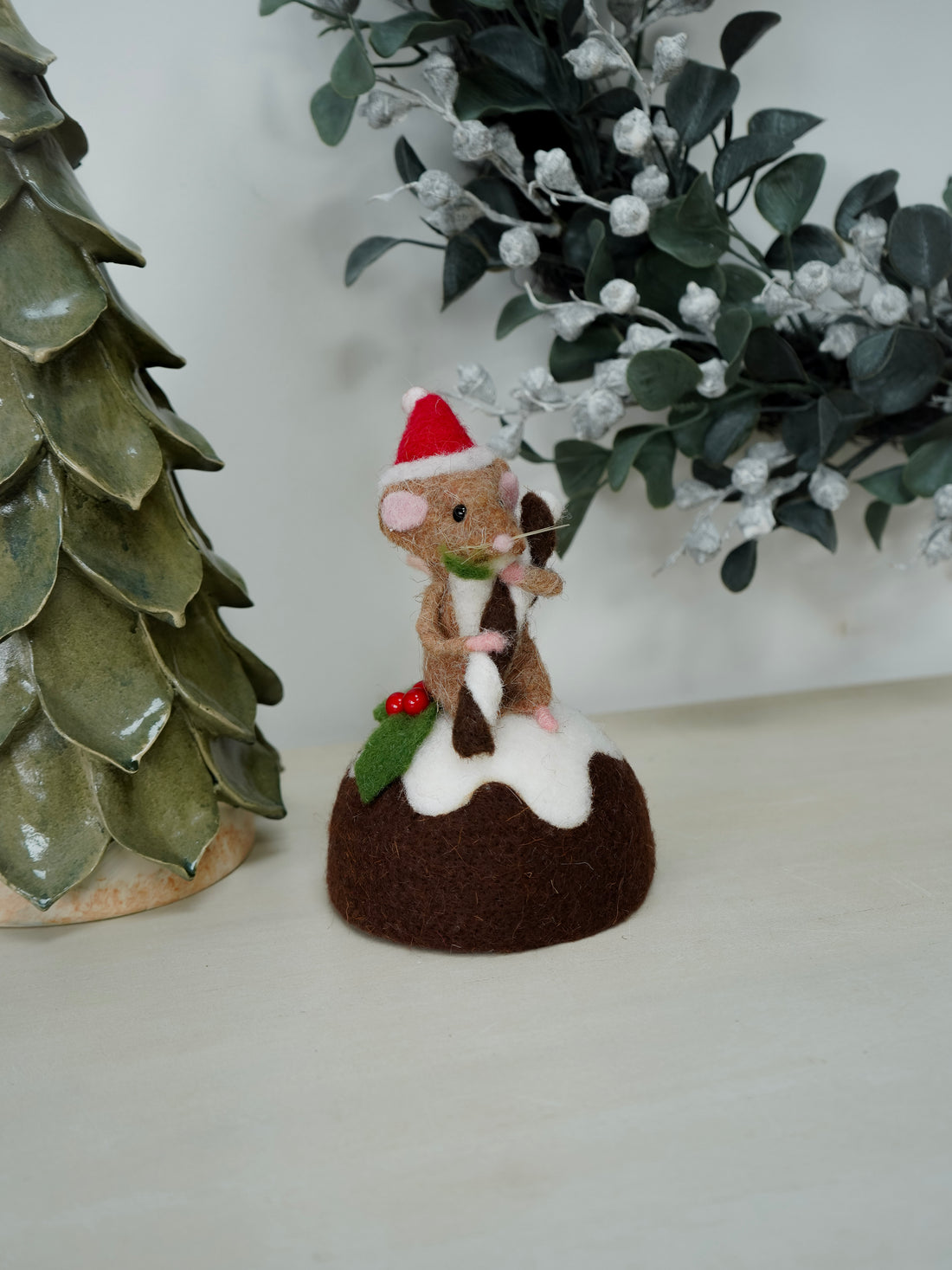 Small decorative figure of a mouse on a Christmas pudding with festive decorations, next to a ceramic tree and greenery.