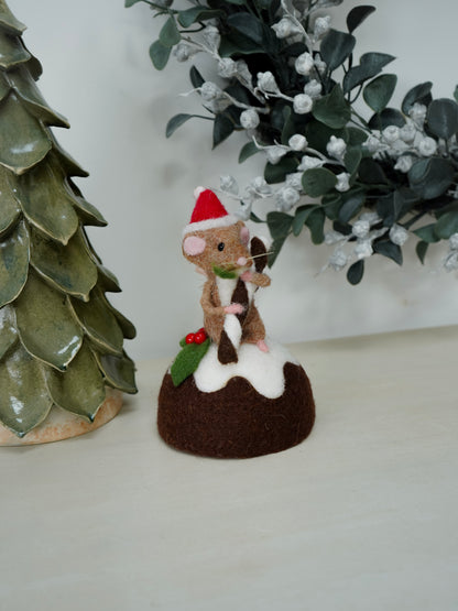 Small decorative figure of a mouse on a Christmas pudding with festive decorations, next to a ceramic tree and greenery.