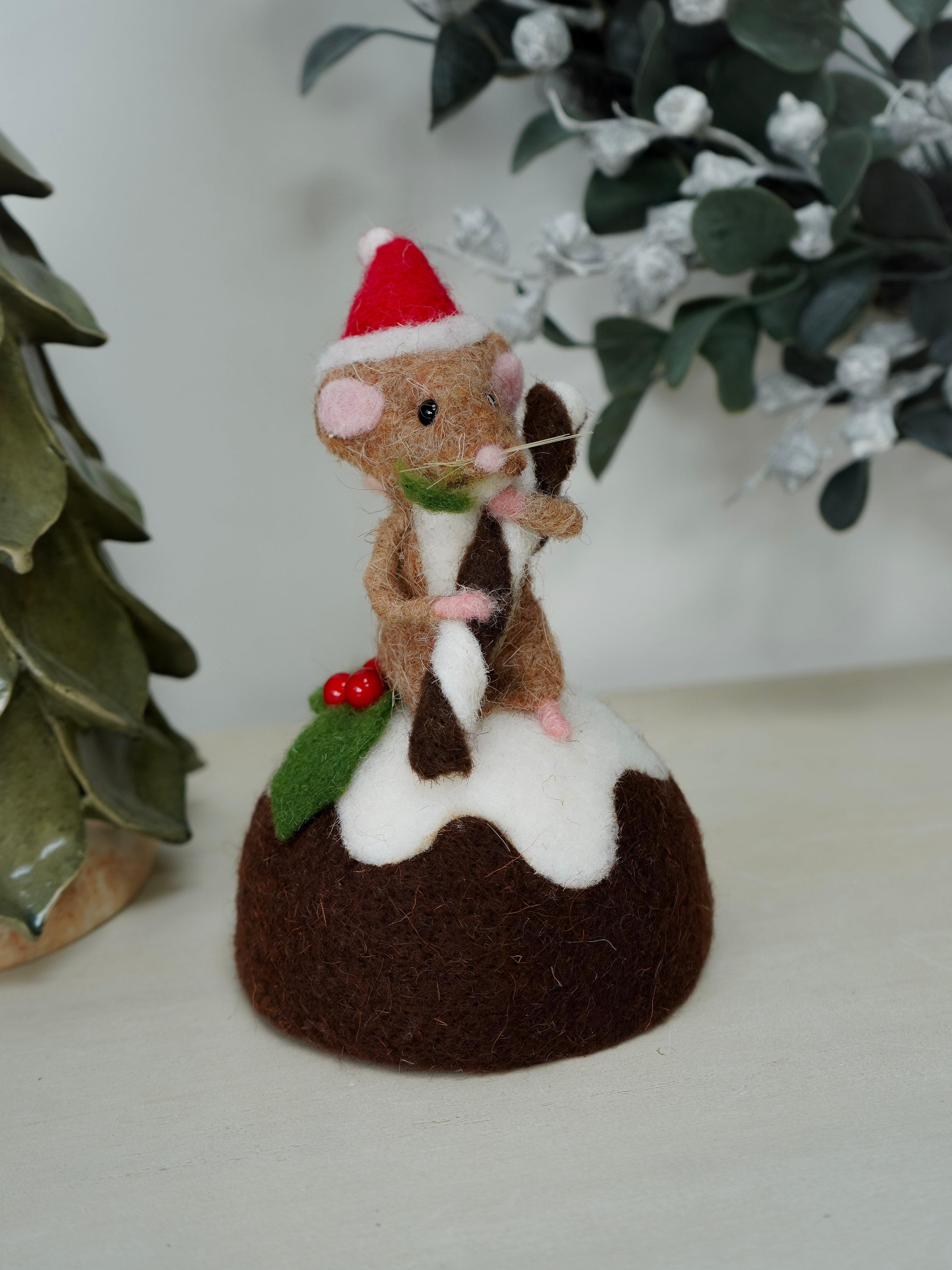 Felted mouse in a Santa hat on a Christmas pudding with mistletoe leaves in the background.