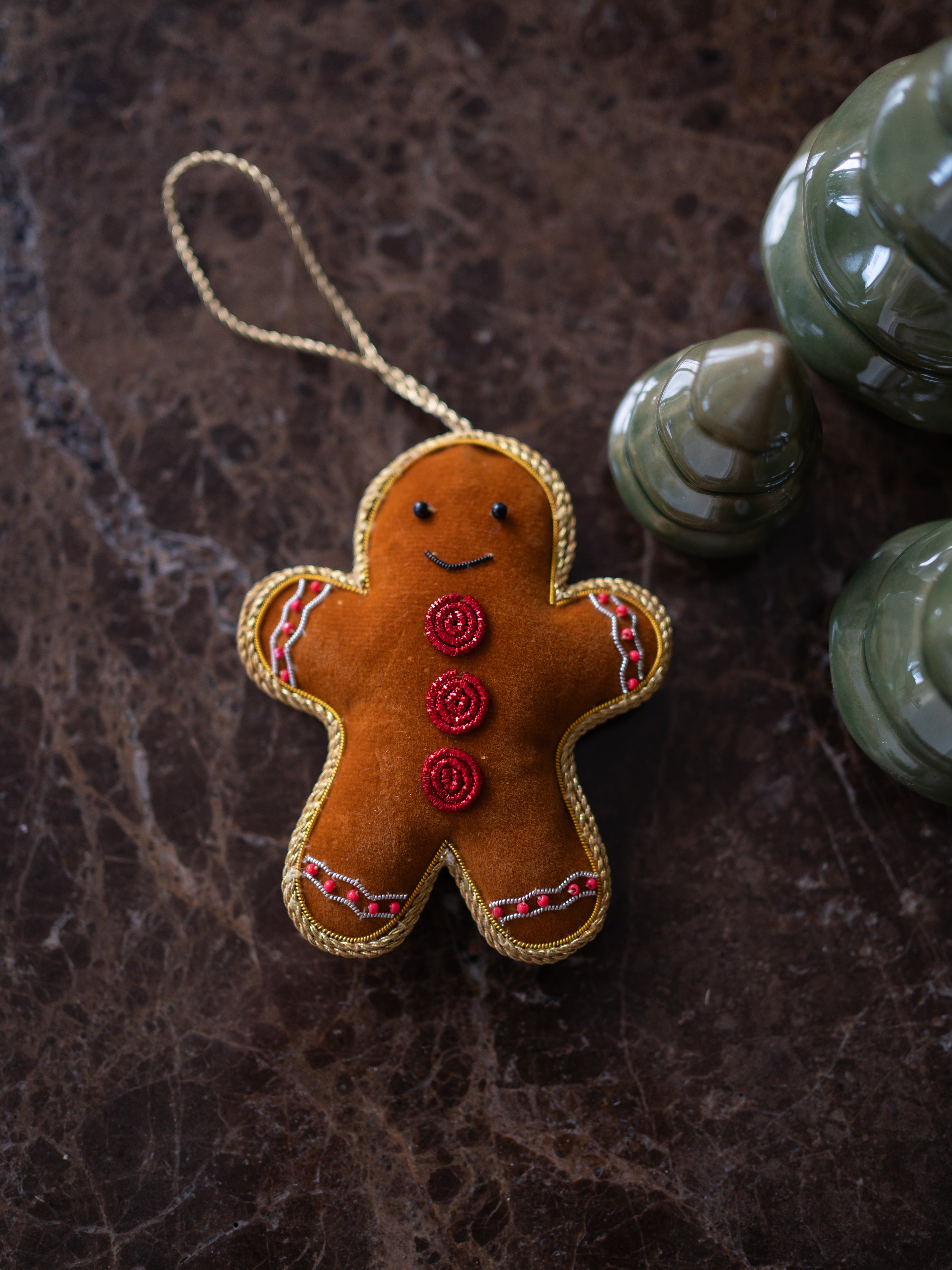 A handmade gingerbread man christmas tree ornament made of brown felt with red embroidered buttons and white/red trim, hanging by a gold cord on a dark brown marble surface with green ceramic ornaments in the background.