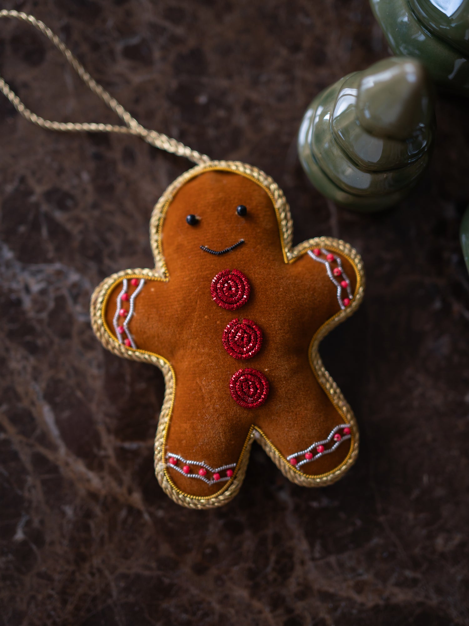 A gingerbread man Christmas tree ornament made of brown felt with gold trim, red button details, and a gold hanging string, photographed on a dark textured surface with a blurred green ceramic object in the background.