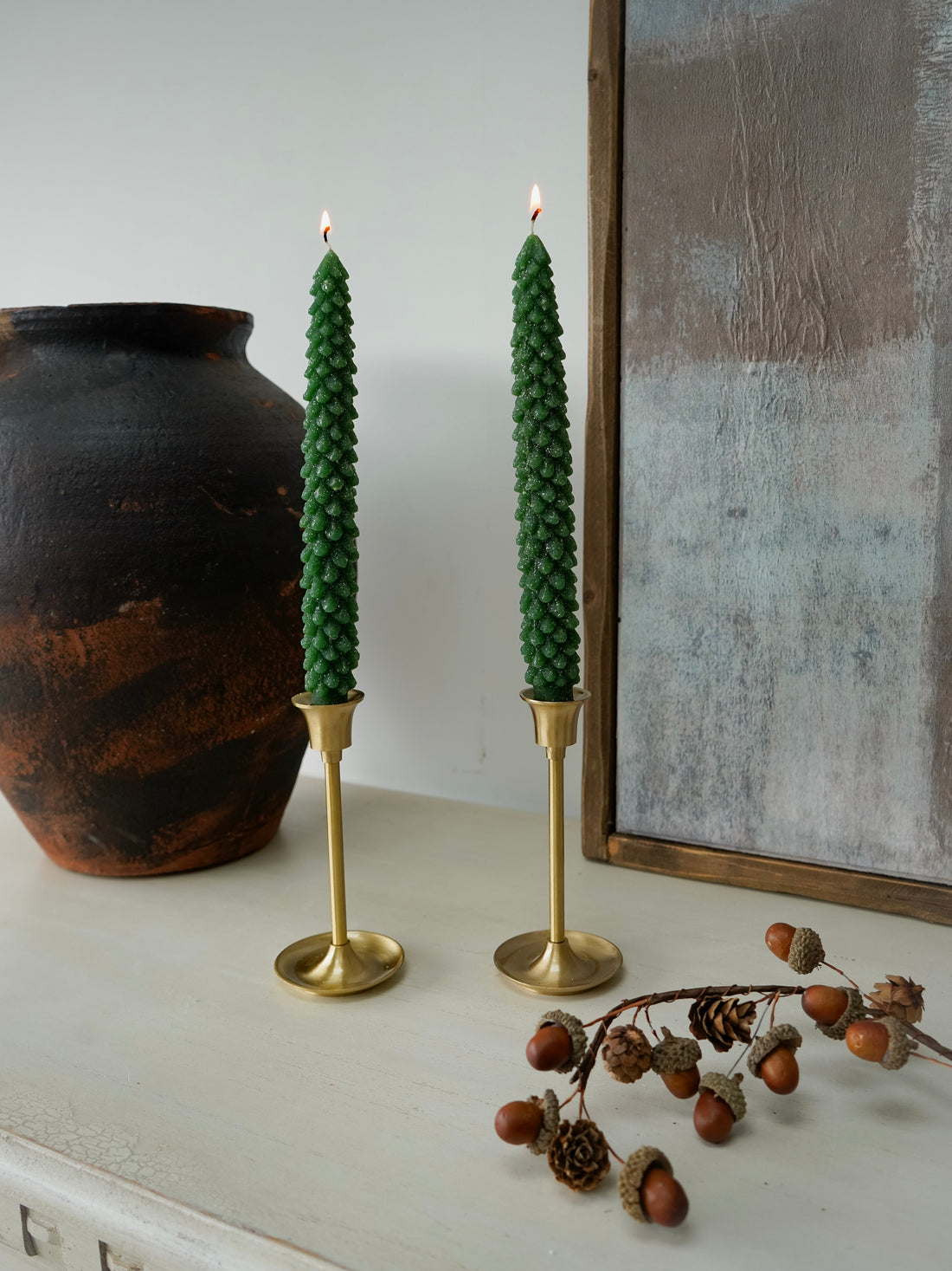 Two green christmas tree dinner candles in gold holders on a white surface with a vase and decorative branch in the background.