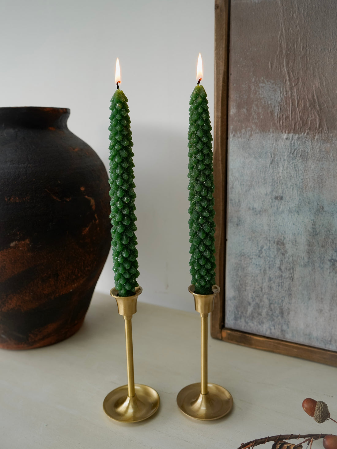 Two green christmas tree dinner candles in gold holders on a neutral surface with a dark vase and framed artwork in the background.
