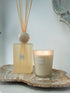 A reed diffuser bottle with beige reeds and a matching lit candle in a frosted glass jar, both labeled &