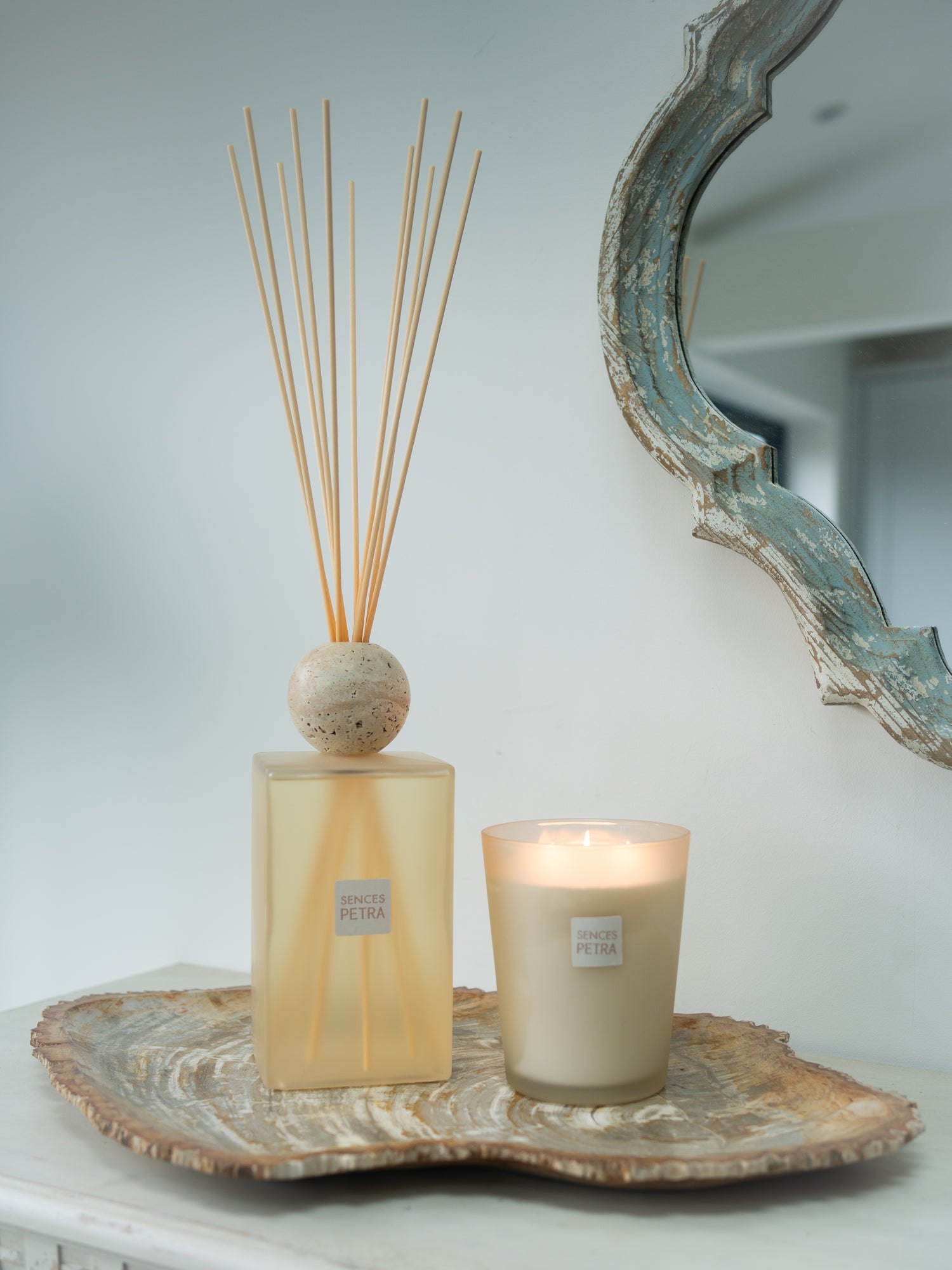 A reed diffuser and a lit candle displayed together on a rustic fossiled wood slice tray against a light gray wall, with part of a distressed blue-green mirror visible.
