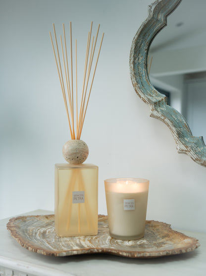 A reed diffuser and a lit candle displayed together on a rustic fossiled wood slice tray against a light gray wall, with part of a distressed blue-green mirror visible.