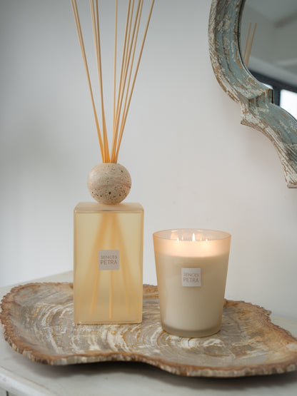 A reed diffuser and a lit candle from the brand SENCES Petra, both with beige labels, styled on a natural wood slice tray against a neutral background.