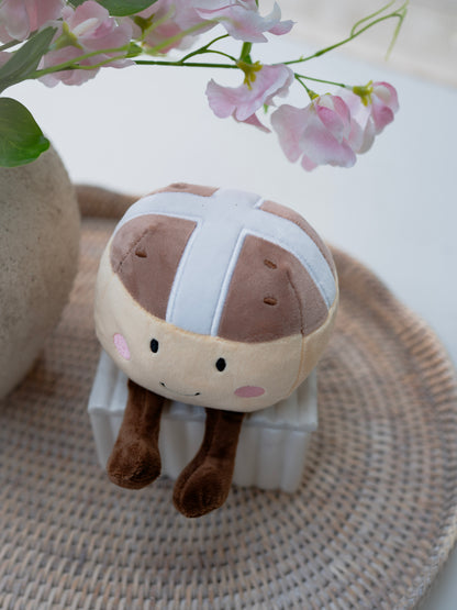 Plush toy resembling a cartoon character with a hot cross bun design on a textured surface with flowers in the background