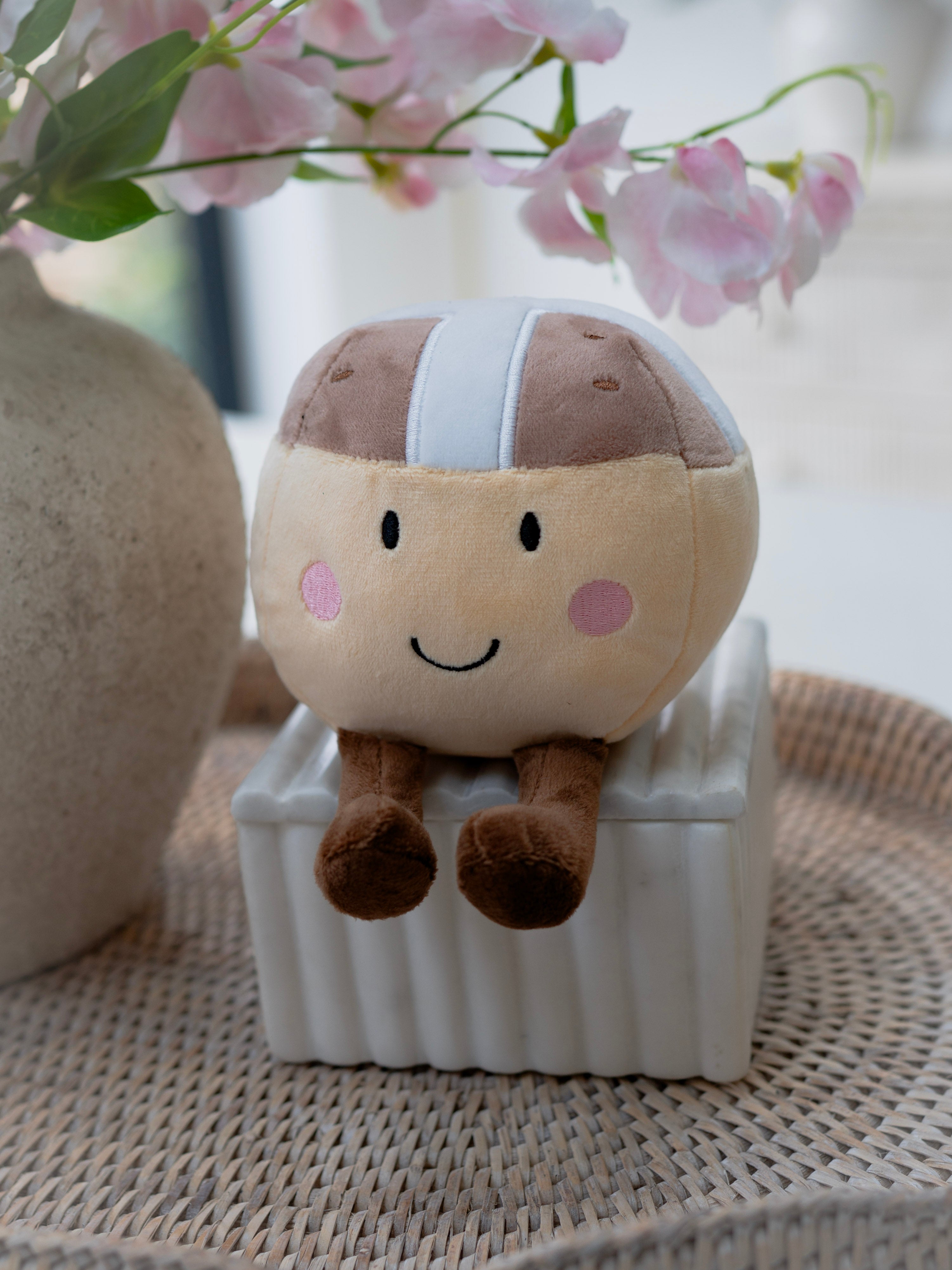 Plush toy resembling a hot cross bun with a smiley face on a white box, surrounded by flowers and a stone-like object.