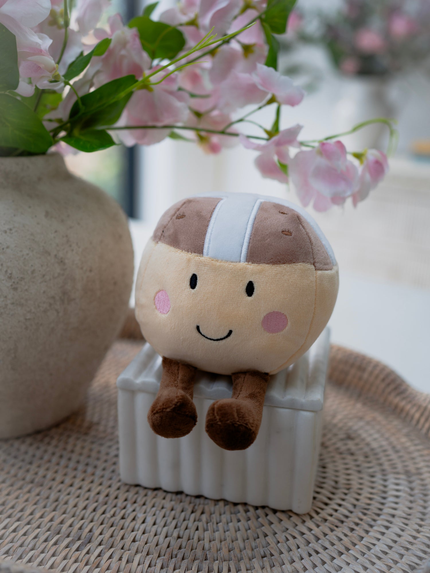Plush toy resembling a hot cross bun sitting on a white block with pink flowers in the background.