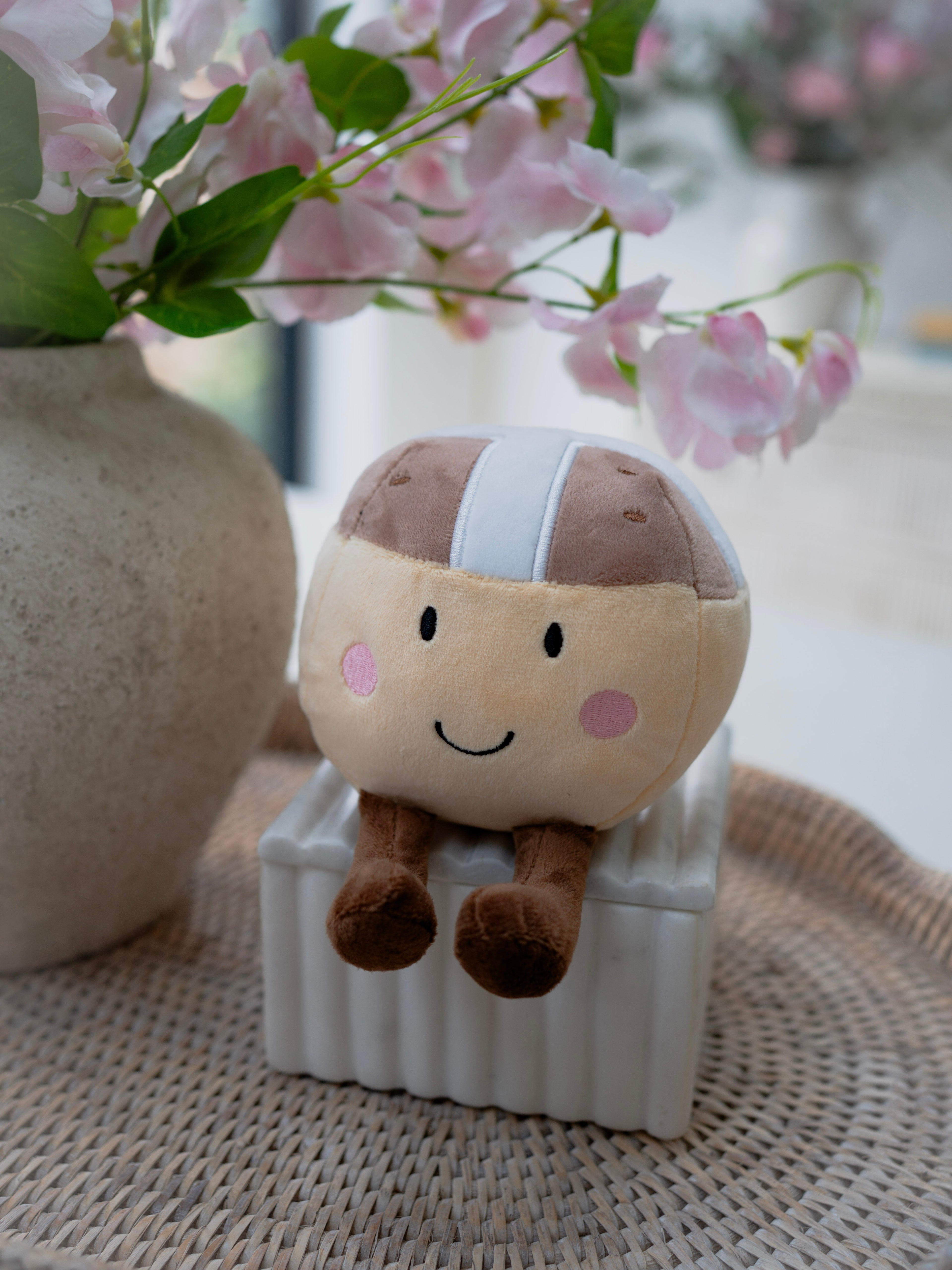 Plush toy resembling a hot cross bun sitting on a white block with pink flowers in the background.