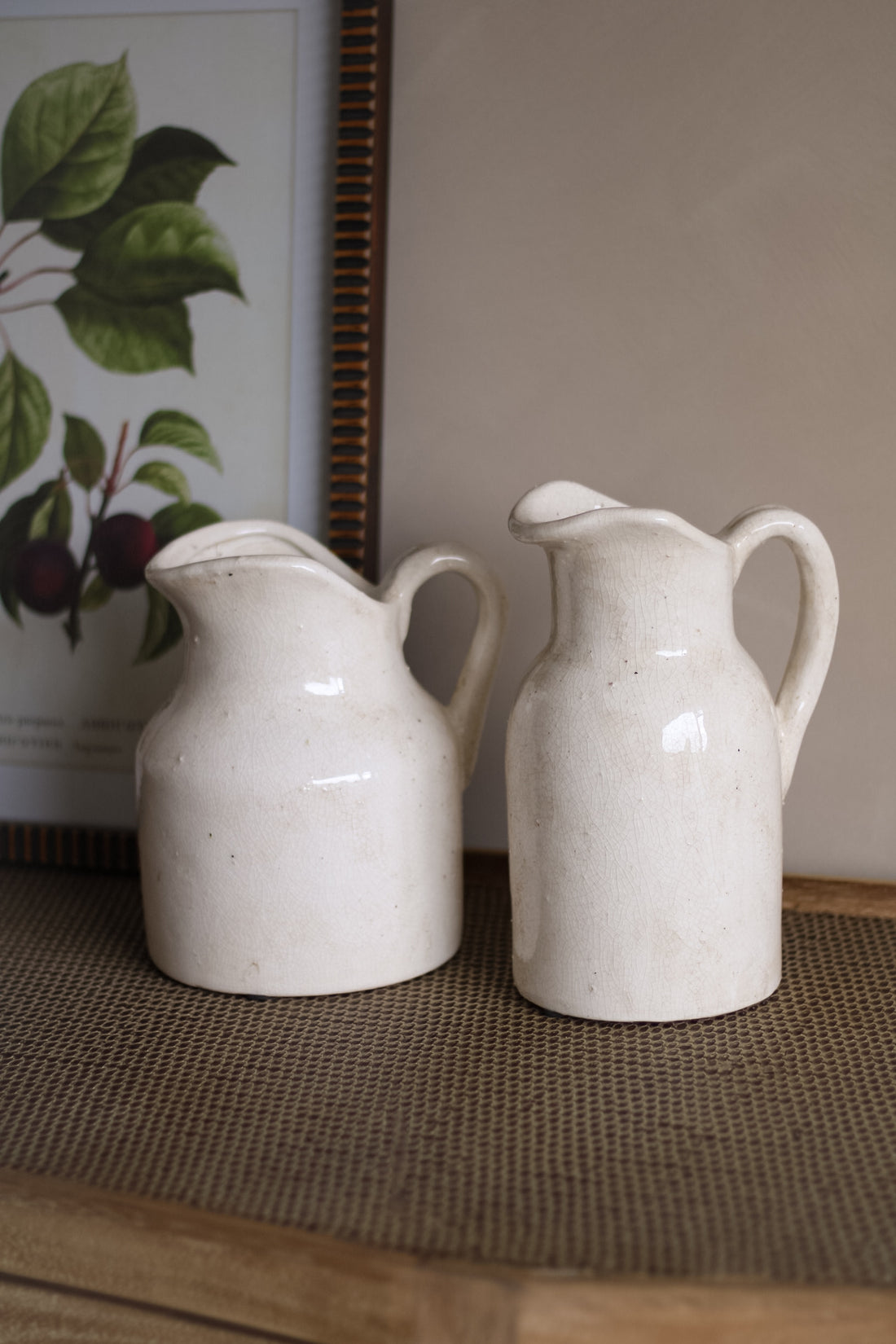 Two white ceramic jugs on a textured surface with a framed picture in the background.
