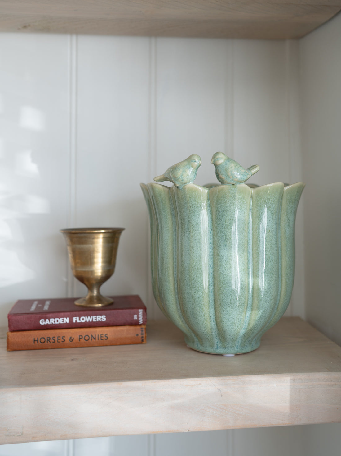 Decorative farmhouse green plant pot with bird figures on a shelf with books and a small cup.