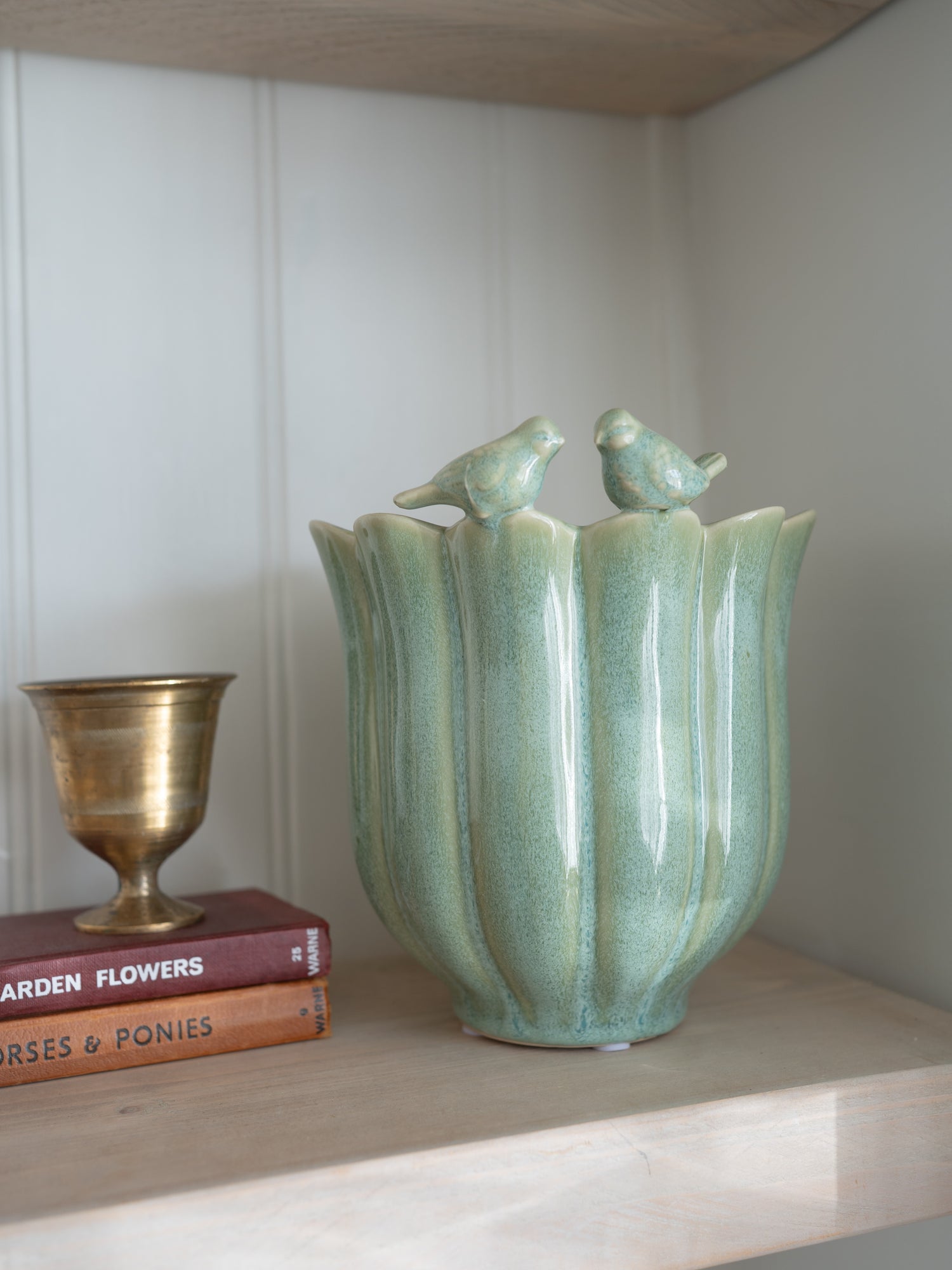 Decorative plant pot in sage green tones with bird designs on a shelf with books and a small cup.