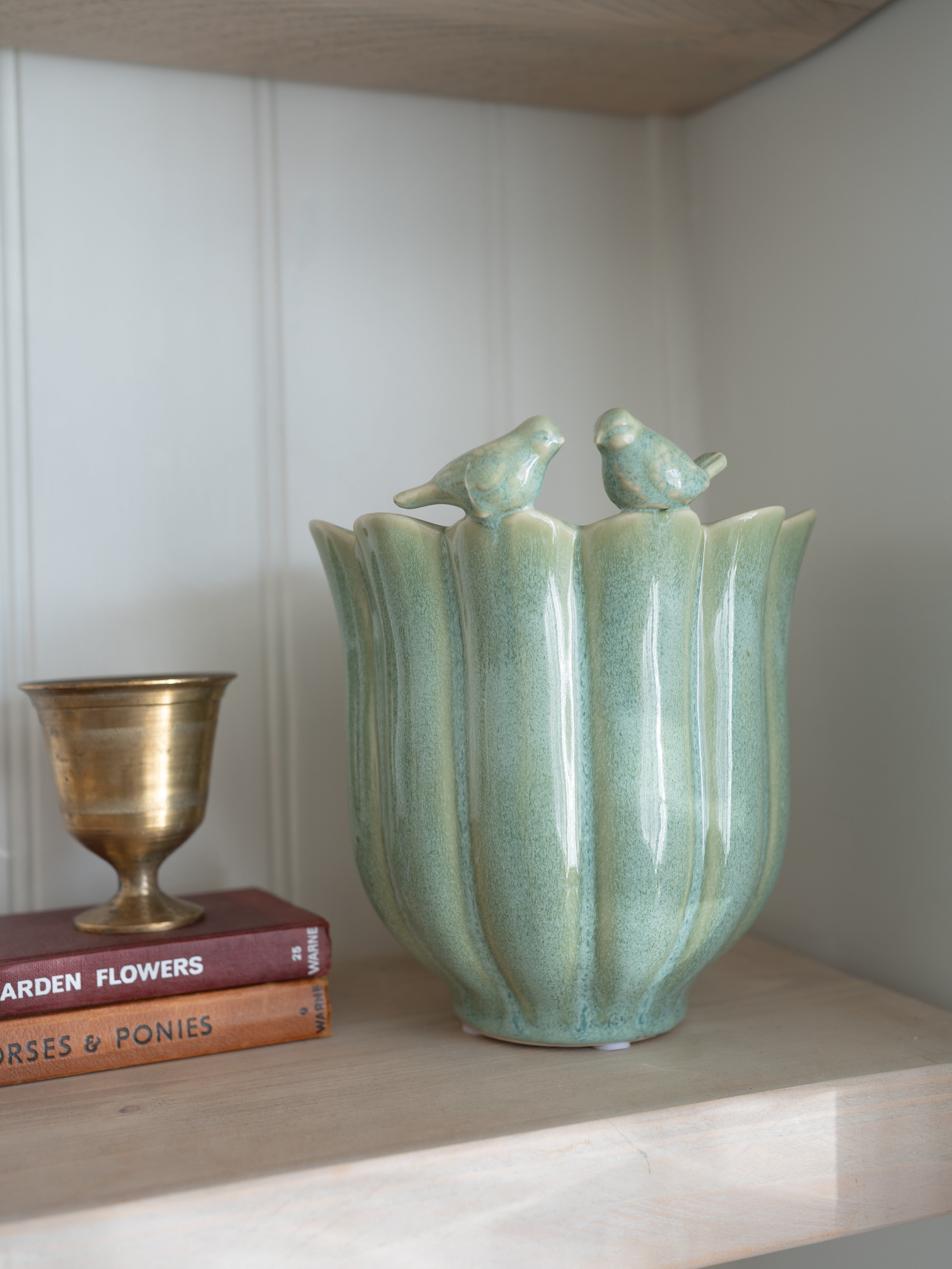 Decorative plant pot in sage green tones with bird designs on a shelf with books and a small cup.
