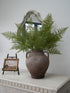 Faux Asparagus Fern sprays in rustic mottled terracotta vase with vintage style wall art of a distant landscape on a beaded small easel