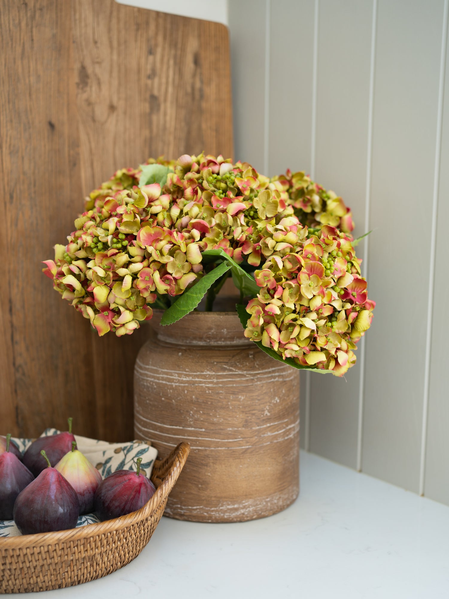 Decorative arrangement with artificial hydrangea flowers in a rustic vase and figs on a woven tray.