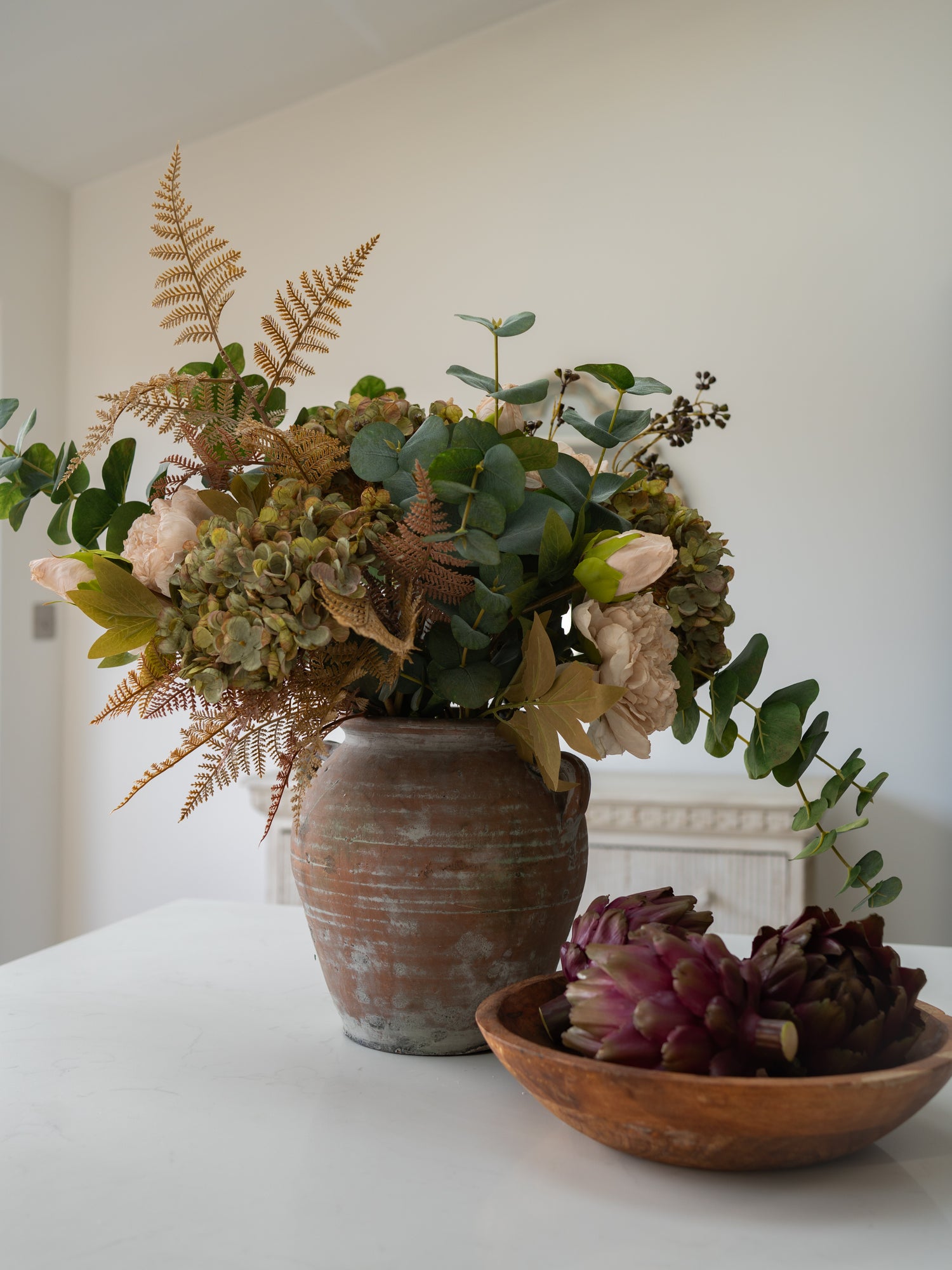 A rustic autumn flower arrangement in a distressed brown ceramic vase, styled on a white table next to a wooden bowl containing purple succulents, against a neutral off-white wall background.