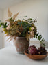 A rustic autumn flower arrangement in a distressed brown ceramic vase, styled on a white table next to a wooden bowl containing purple succulents, against a neutral off-white wall background.