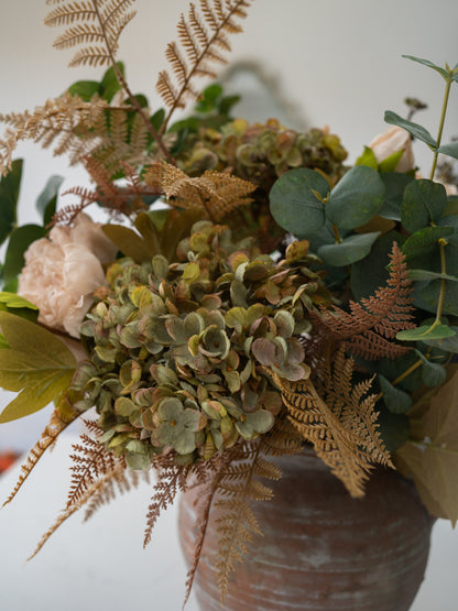 A rustic autumn floral arrangement in a textured brown ceramic pot, featuring muted green hydrangeas, tan ferns, eucalyptus leaves, and pale blush pink flowers against a soft off-white background.