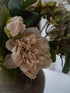 A close-up of a autumn floral arrangement featuring a large cream peony, a pale blush bud, and muted green hydrangea blooms in a brown ceramic vase.