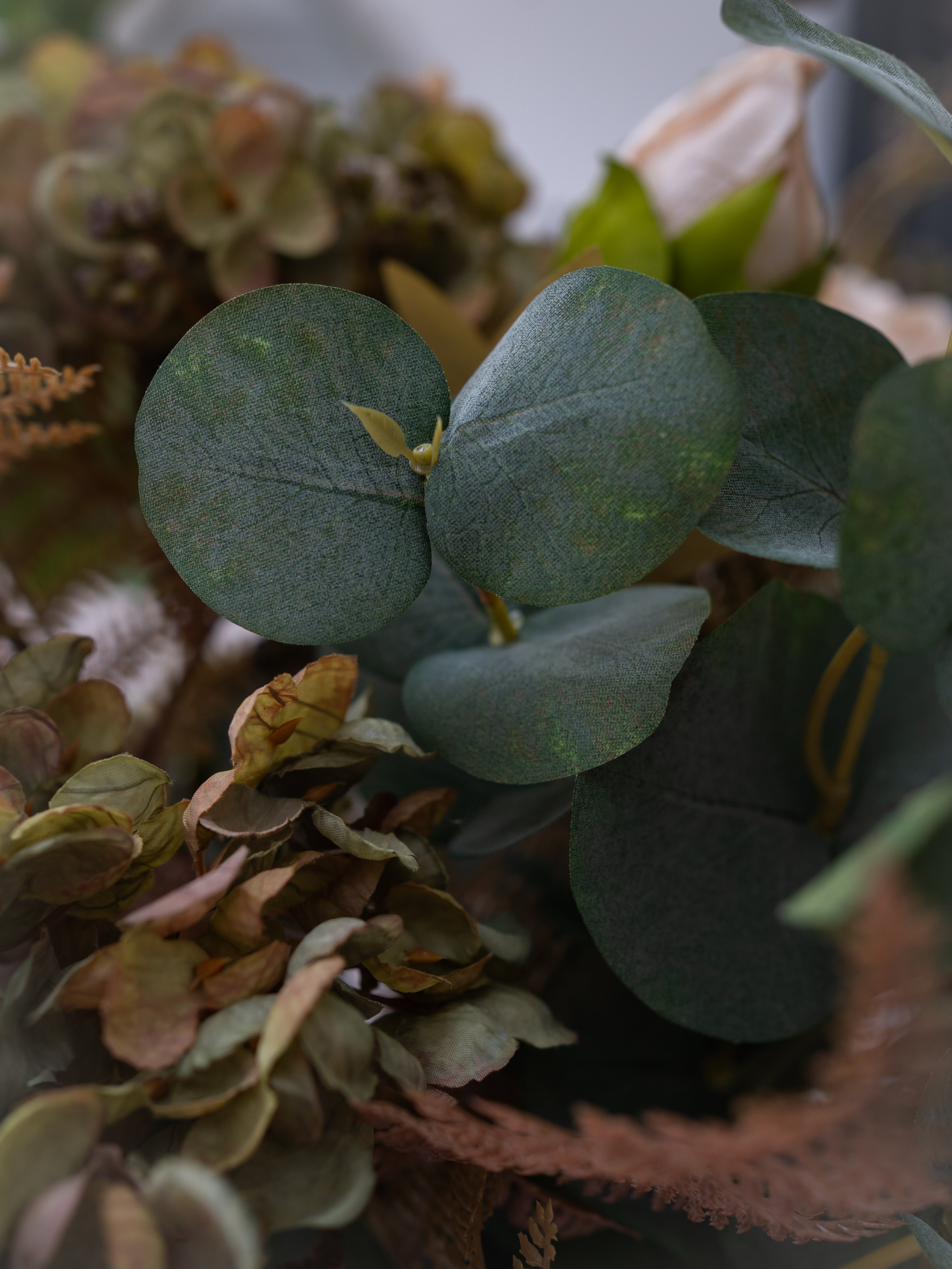 Close-up of a decorative faux floral arrangement featuring round dark green eucalyptus leaves alongside dried muted flowers in tan, brown, and olive tones, with a soft blurred background.