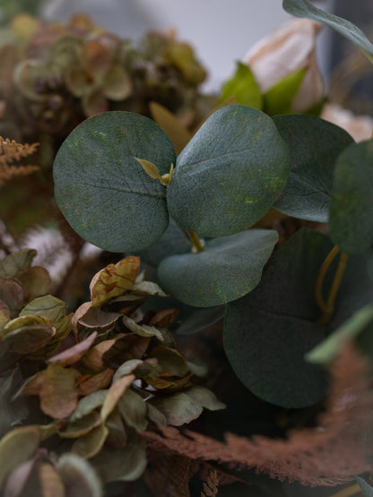 Close-up of a decorative faux floral arrangement featuring round dark green eucalyptus leaves alongside dried muted flowers in tan, brown, and olive tones, with a soft blurred background.