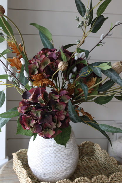 White textured vase with a bouquet of autumn flowers and greenery on a woven mat.