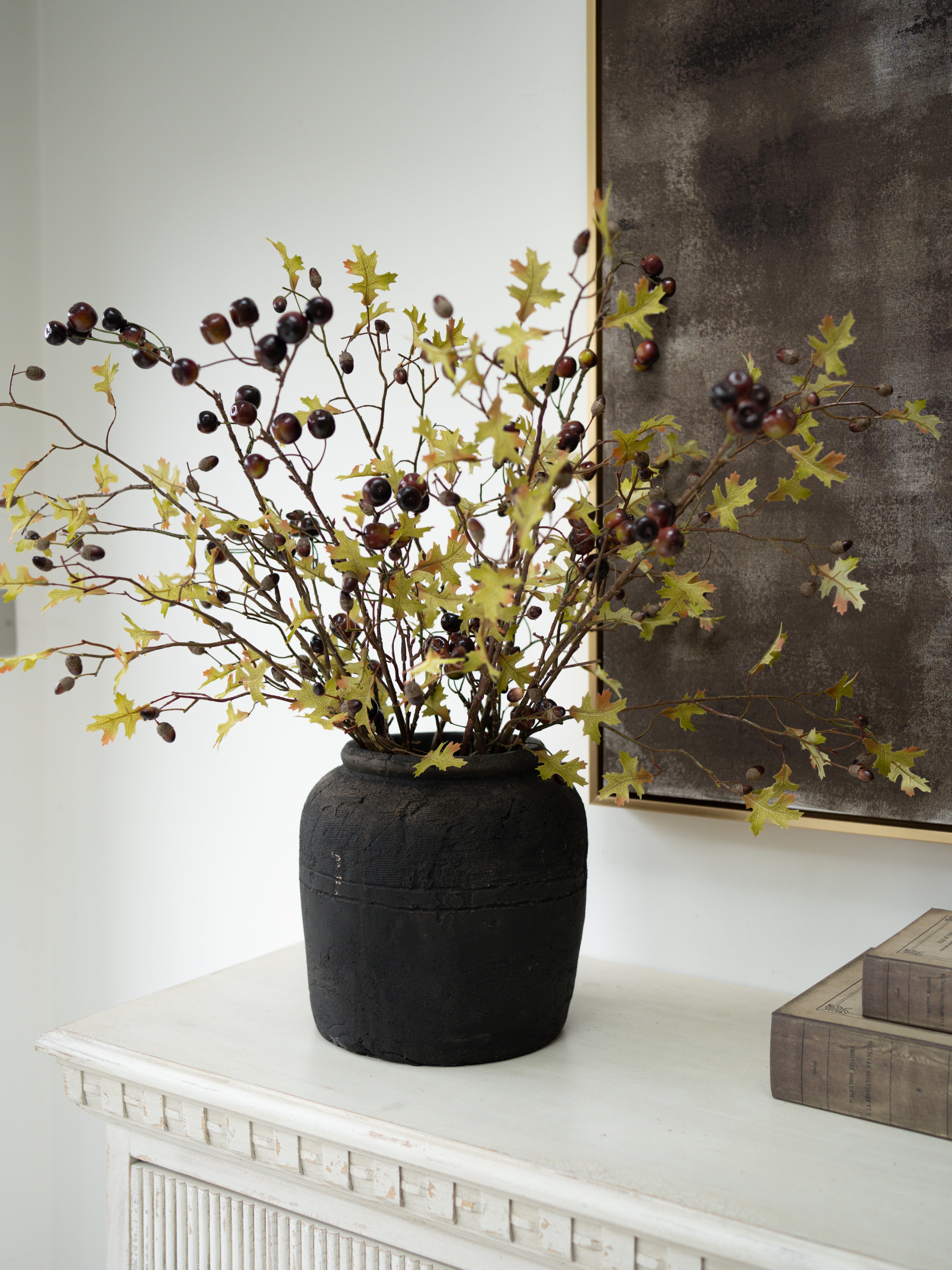 Faux autumn oak branch arrangement with yellow-green leaves and dark acorns in a dark textured ceramic vase, displayed on a white mantelpiece against a white wall.