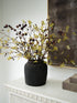 Faux autumn oak branch arrangement with yellow-green leaves and dark acorns in a dark textured ceramic vase, displayed on a white mantelpiece against a white wall.