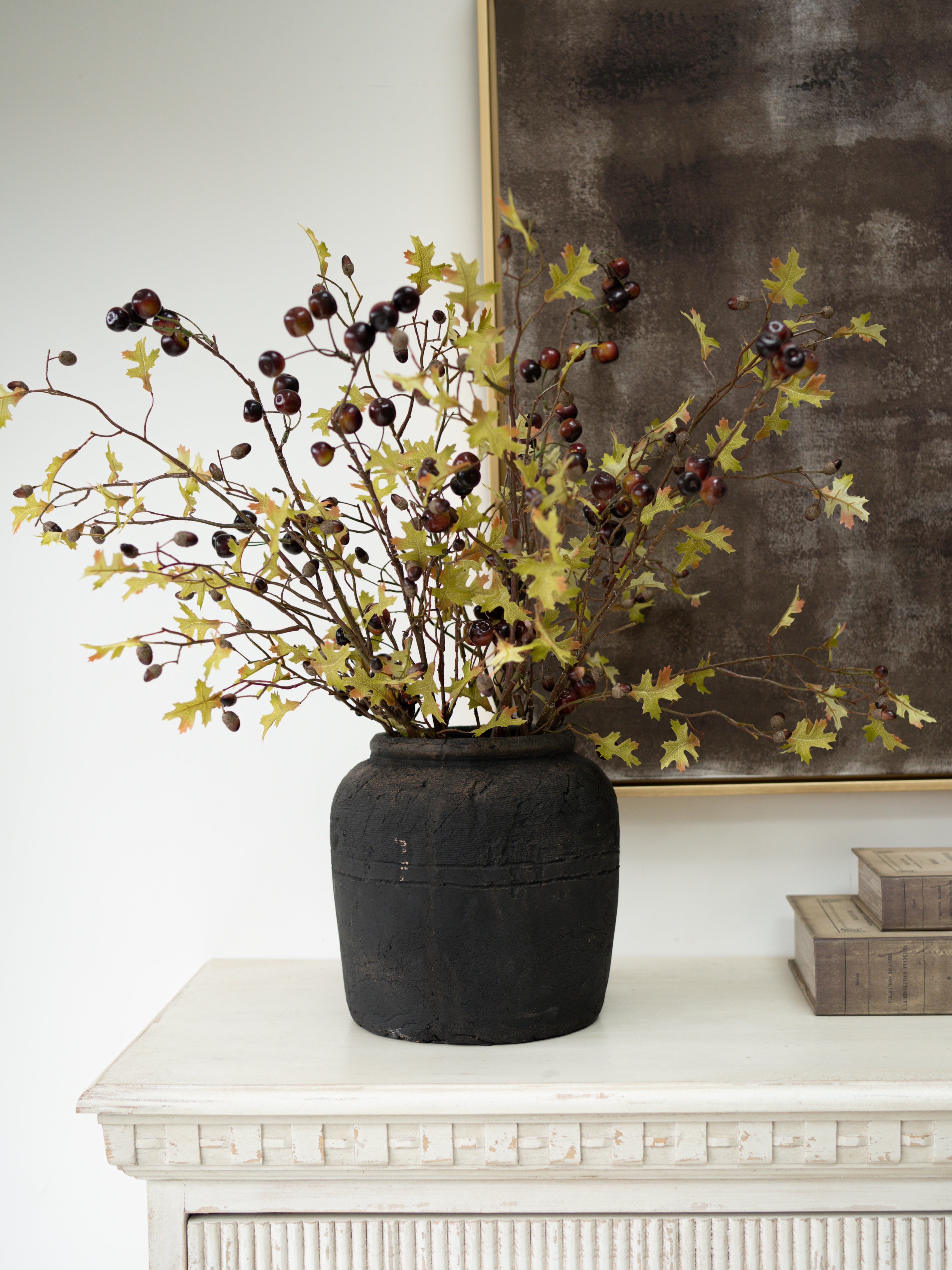 Autumn artificial floral arrangement with yellow-green oak leaves and dark wild cherries in a dark textured ceramic pot, displayed on a white mantelpiece against a white wall and dark framed artwork.
