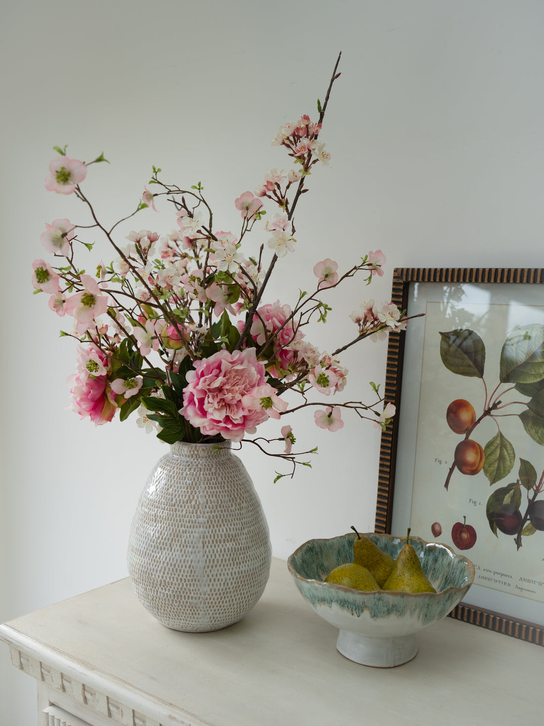 Vase with pink flowers on a table next to a bowl of pears and a framed picture.