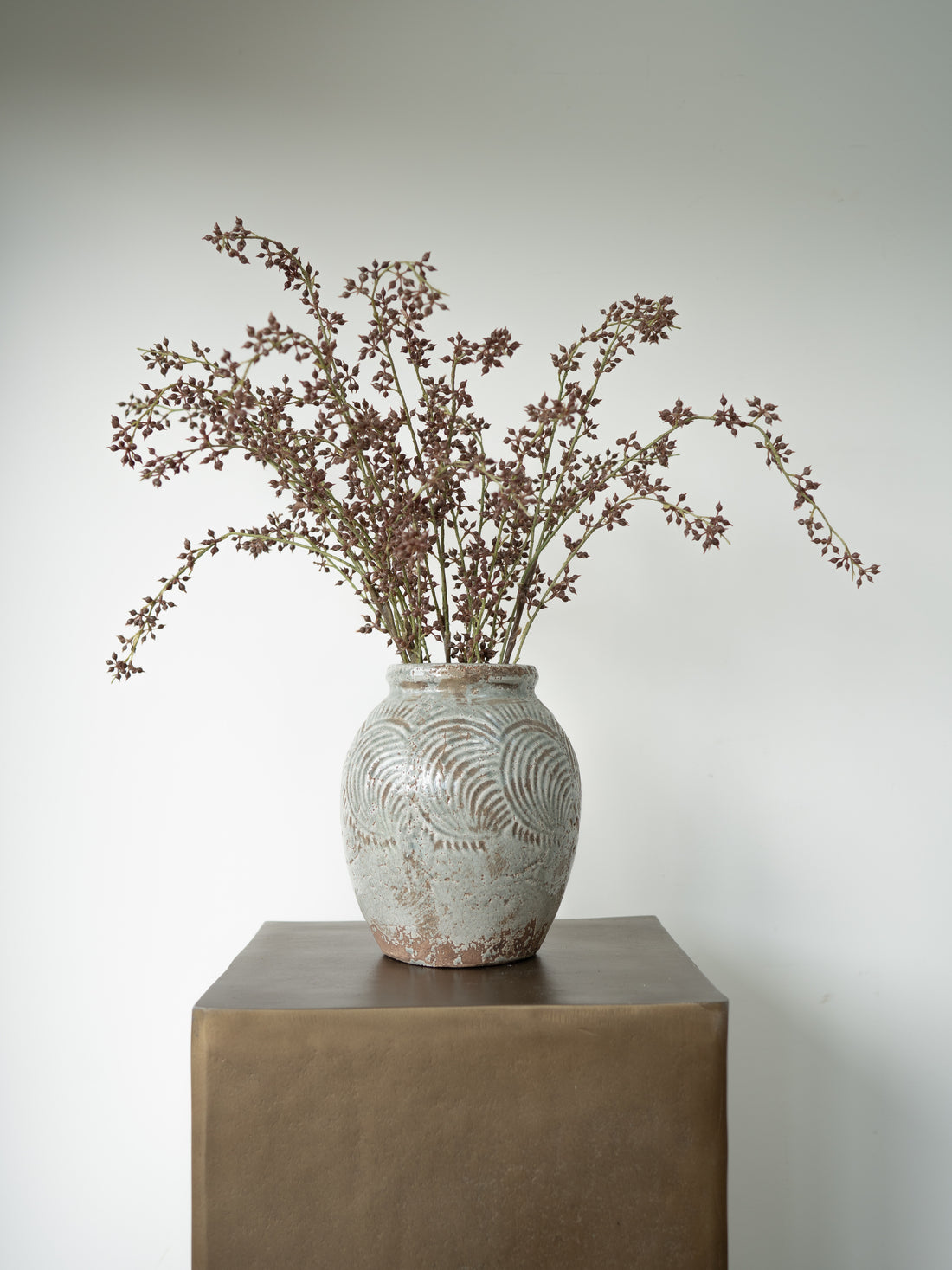 A rustic ceramic vase with a textured, distressed finish sits on a dark brown pedestal, holding a bouquet of dried brown branches against a plain light gray background.