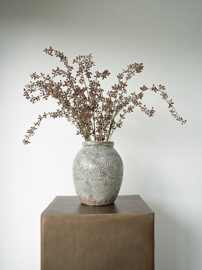 A rustic ceramic vase with a textured, distressed finish sits on a dark brown pedestal, holding a bouquet of dried brown branches against a plain light gray background.