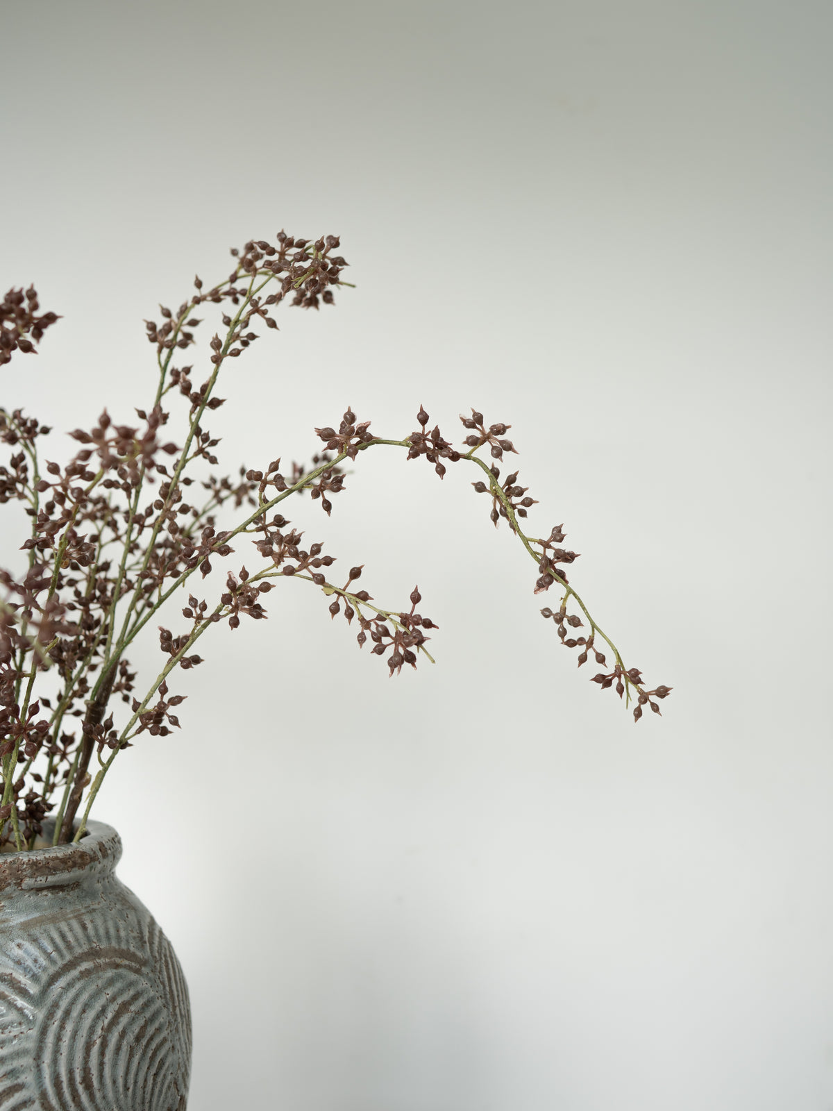 Dried brown flower stems arranged in a textured gray ceramic vase against a plain light gray background.
