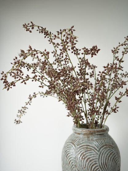 Dried brown flower stems arranged in a textured gray ceramic vase against a plain light gray background.