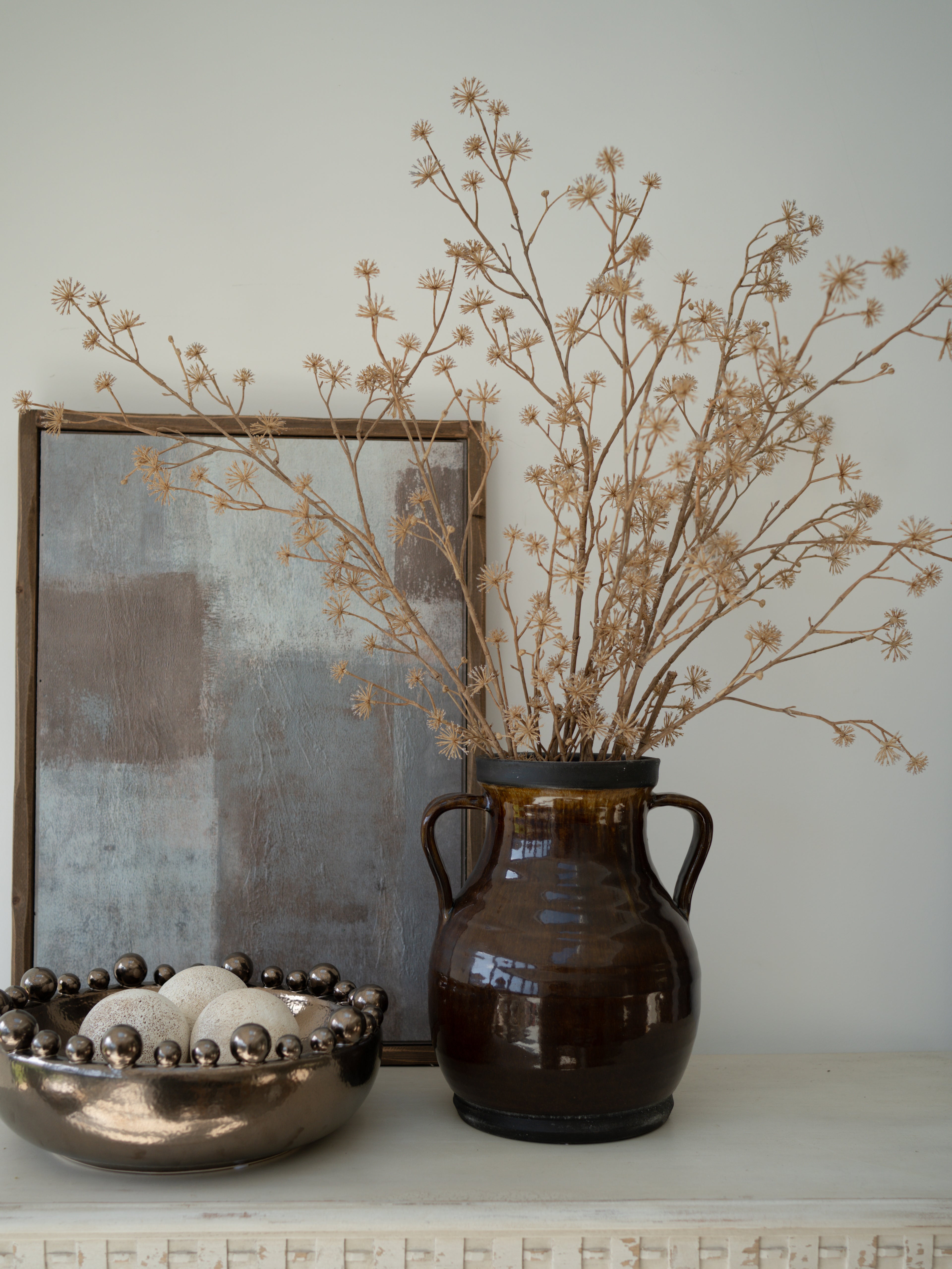 Decorative setup with a brown vase, faux brown dogwood branches, and a framed abstract painting on a shelf.