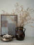 Decorative setup with a brown vase, faux brown dogwood branches, and a framed abstract painting on a shelf.