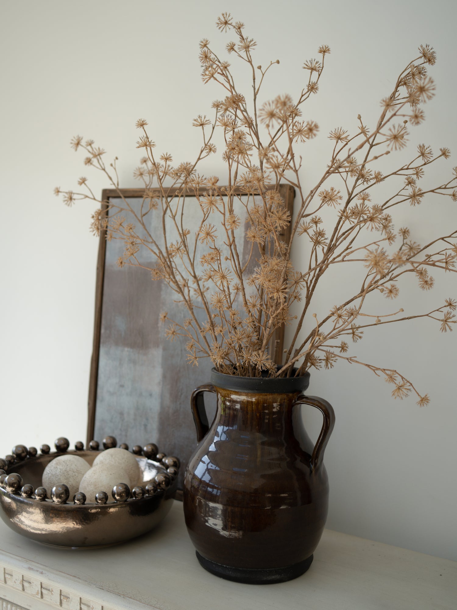 Brown ceramic vase with faux brown dogwood branches on a surface, with a blurred mirror in the background.