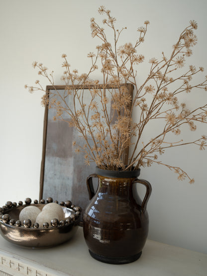 Brown ceramic vase with faux brown dogwood branches on a surface, with a blurred mirror in the background.